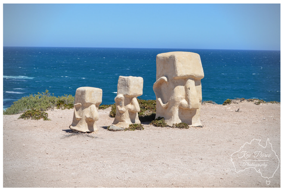 Three beige, blocky statues, part of the Elliston coastal art trail, stand facing the deep blue ocean under a clear blue sky.

The smallest is on the left, and the largest is on the right, which features a trunk and ear detailing. The statues are positioned on a sandy, scrub covered cliff edge overlooking the crashing waves.