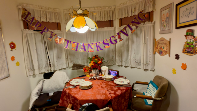 A Thanksgiving table for two set with a bronze tablecloth, fine china, two cranberry sauces, pumpkin and leaf decorations, and an autumnal flower arrangement. A Happy Thanksgiving banner in purple and gold hangs over the white drapes. Leaves are pasted to the wall. 
 
