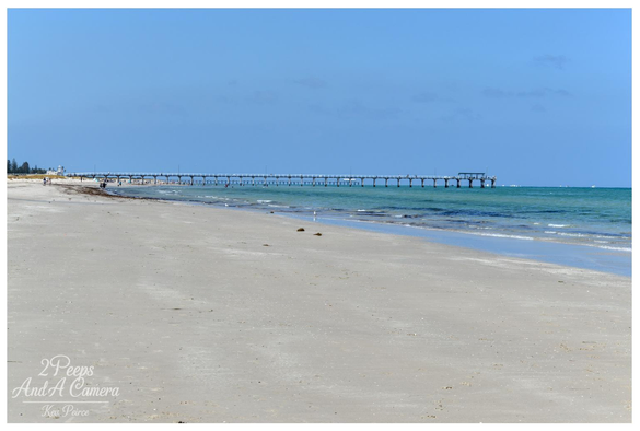 A tranquil colour photograph capturing the wide, sandy beach and the historic Semaphore Pier in Adelaide, South Australia.

The long, horizontal pier extends over the turquoise water of Gulf St Vincent into the distance under a vast, cloudless light blue sky.

The foreground is dominated by the pale, sun-drenched sand, with the water gently lapping the shore.
