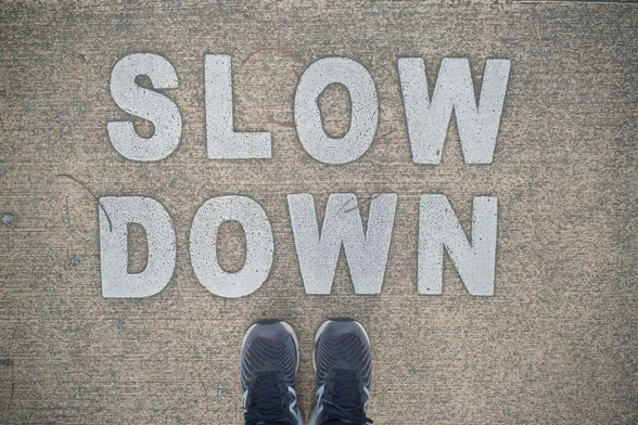 Close-up photo of a concrete footpath with a white-coloured 'slow down' warning painted on it. The front of the photographer's black-and-white sneakers are visible in the bottom of the frame.