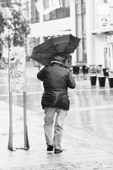 Das Foto zeigt eine Straßenszene in Schwarz-Weiß in der St. Pöltenter Innenstadt an einen regnerischen windigen Tag. Man sieht eine Person mit Regenschirm der durch den Wind verbogen ist.