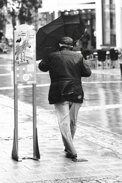 Das Foto zeigt eine Straßenszene in Schwarz-Weiß in der St. Pöltenter Innenstadt an einen regnerischen windigen Tag. Man sieht eine Person mit Regenschirm der durch den Wind verbogen ist.