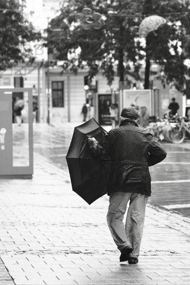 Das Foto zeigt eine Straßenszene in Schwarz-Weiß in der St. Pöltenter Innenstadt an einen regnerischen windigen Tag. Man sieht eine Person mit Regenschirm der durch den Wind verbogen ist.