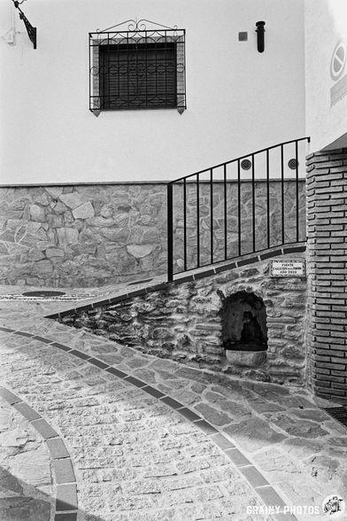 A winding stone pathway leads up to a small fuente inset into a textured stone wall with a black iron railing. Above, a decorative window with burglar bars is set against a white wall, while a small fountain can be seen near the base of the slope.