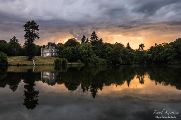 Photographie en couleur présentant une vue partielle du parc d'un château, en fin de journée, sous un ciel orageux.
La photographie a été prise depuis le bord d'un étang à la surface duquel se reflètent les arbres de la rive opposée et notamment, un magnifique séquoia sur la gauche du cadrage. À droite de celui-ci, on peut voir la façade d'un château du XVIIIe siècle.
Le ciel est encombré de gros nuages, menaçants, très gris, mais se "dorant" à proximité de l'horizon et du soleil couchant.
Bien évidemment, ciel, végétation et château se reflètent dans le beau miroir de la surface de l'étang.