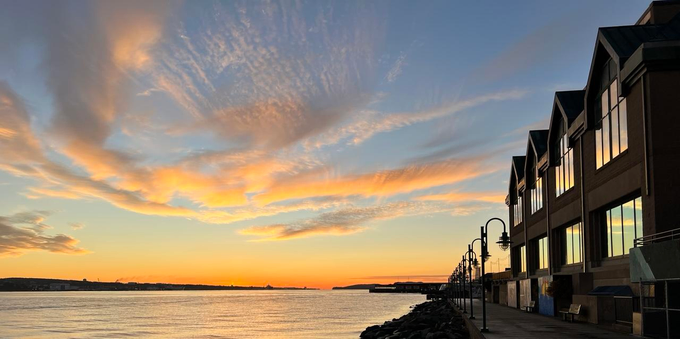 A colourful sunrise sky reflects from windows along a harbour side boardwalk. The two-storybuilding is on the right of image, with peaked gables over four sets of windows. The boardwalk is lined with streetlights. The remainder of the foreground is an expanse of open harbour with a thin strip of black land along the horizon. The water shows muted versions of the blues, yellows and oranges in the sky that is streaked with  thin wispy clouds. [Halifax (NS, Canada) November 2025]