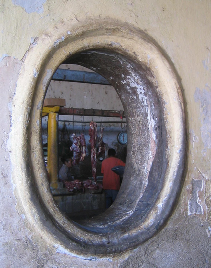 Der Zugang zum Fleischmarkt blieb uns verwehrt. Den Grund hierfür können wir mit dem Blick durch dieses Fenster nur erahnen. Es ist ein ehemals weißes, aber nun sehr abgegriffenes ovales Luftloch in der Mauer zwischen den Markthallen. In der Fleisch-Abteilung hängen die angepriesenen Stücke am Haken. Ein Kühlschrank ist nirgends zu sehen. 