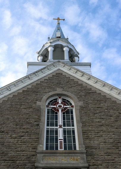 Photograph of a window on the front of a stone church, taken from the foot of the church and looking upwards, with a white wooden cross and red decorations in the centre of the frame, and below it a stone inscribed "Erigee AD 1884". Above is the church steeple, with a golden cross at the top where a black crow is perched. The sky is blue with white fluffy clouds.

Photographie d'une fenêtre sur le devant d'une église en pierres, prise au pied de l'église et regardant vers le haut, avec une croix blanche en bois et des décorations rouges en plein milieu du cadre, et et en dessous une pierre où est écrit "Erigee AD 1884". Au-dessus se trouve le clocher de l'église, avec une croix dorée au sommet où est perchée une corneille noire. Le ciel est bleu avec de petits nuages blancs.