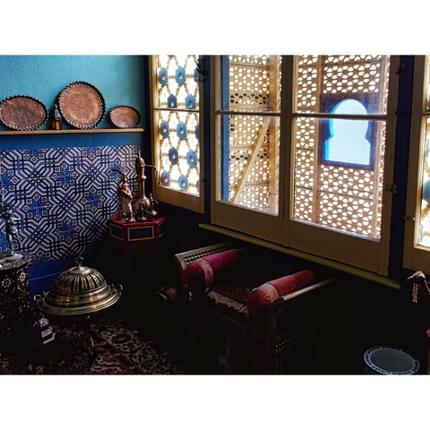 A cozy room with Moroccan decor, featuring ornate blue tiles, brass trays, and wooden latticework. Warm light filters through patterned windows.