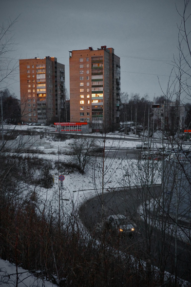 Morning. Cityscape, view from a hill. The sky will be covered with clouds. There are bushes in the foreground. In the middle plan there is a building and a passing car with burning headlights. In the background there are two high-rise, red buildings with “burning” windows from the glare of the sun.