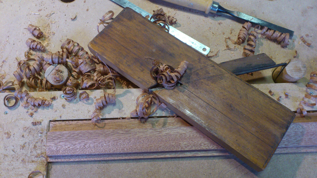 Photo d'un guillaume en bois, posé sur le flan. C'est un rabot très fin, un peu comme une planche.
On voit un réglet et un ciseaux à bois également, ainsi que des copeaux et une petite planche rabotée. 