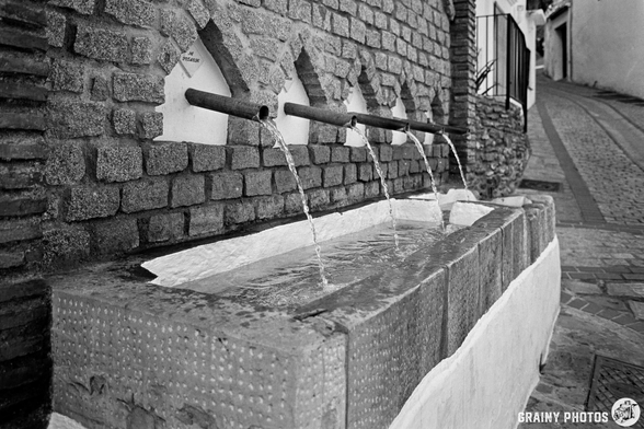 An old stone fountain (fuente) with five spouts, water flowing gently from each, set against a textured brick wall in a narrow street. The scene is captured in black and white, highlighting architectural details and the calming sound of water.