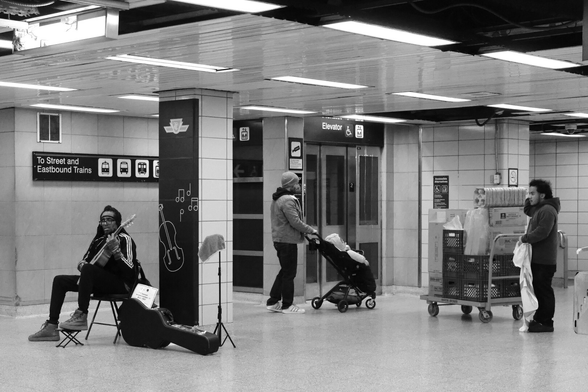 The concourse level between the trains and the street. At right, a young man stands with a trolley full of supplies for one of the businesses. Centre, in front of the elevator, a man pushes a stroller. At left, sitting by one of the pillars, an older man with dreadlocks holds a guitar, the case at his feet.