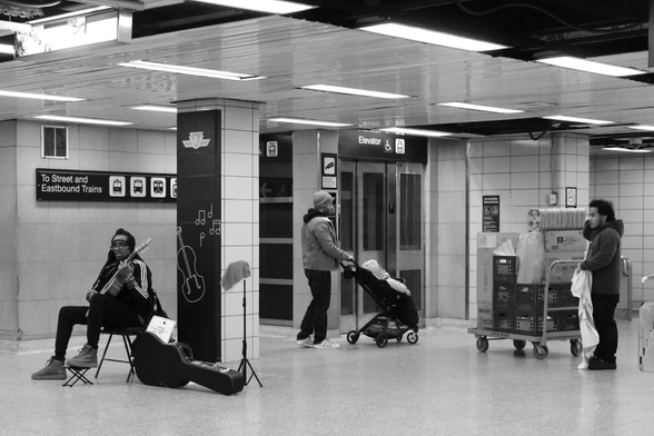 The concourse level between the trains and the street. At right, a young man stands with a trolley full of supplies for one of the businesses. Centre, in front of the elevator, a man pushes a stroller. At left, sitting by one of the pillars, an older man with dreadlocks holds a guitar, the case at his feet.