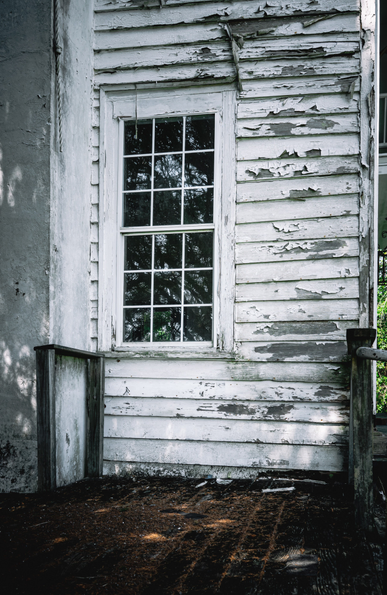 The exterior of an abandoned or severely deteriorated wooden building with white painted horizontal siding that is extensively peeling and flaking off, revealing weathered gray wood underneath. A tall, narrow double-hung window with white painted frame and multiple panes reflects trees in its glass. The window is flanked by portions of what appears to be corner trim or adjacent wall sections. At the bottom of the frame, dark wooden posts or supports are visible, along with a deteriorated wooden floor or porch covered in dirt, debris, and decomposing organic matter. The overall condition suggests long-term neglect and exposure to the elements.