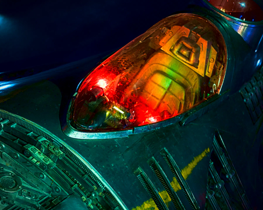 Blick durch das rot erleuchtete Fenster in das Cockpit eines Raumjägers aus der Filmreihe Star Wars. Man schaut von vorn auf den Pilotensitz. Die Metallhaut des kleinen Raumjägers zeigt verschiedene Einlässe, Kabel und andere Gegenstände. 
Ein schmaler, gelber Streifen wie mit einem alten Pinsel gezogen zieht sich über die Seite des Raumjägers. 
. 
View through the red-lit window into the cockpit of a starfighter from the Star Wars movie series. You are looking at the pilot's seat from the front. The metal skin of the small starfighter shows various inlets, cables and other objects.
A narrow yellow stripe, as if drawn with an old paintbrush, runs across the side of the starfighter.
. 
