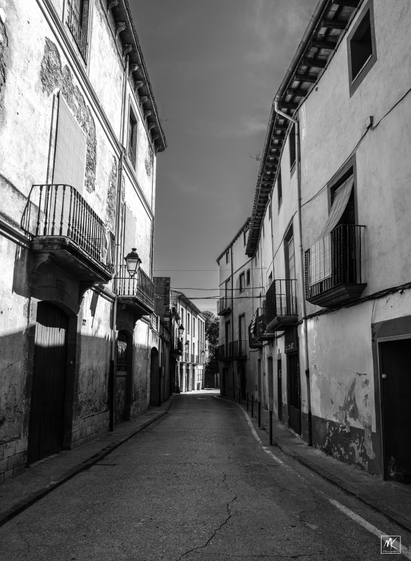 Black and white photo looking down an empty street lined with houses in an old Catalonian town. 