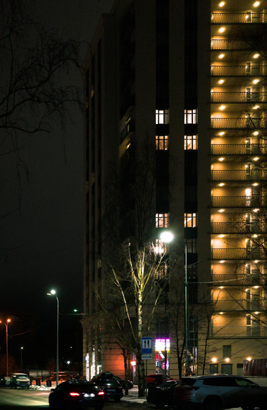 The photo shows a high-rise building with a vertical line of illuminated balconies. There are also lights on in several windows. In front of the house there is a road along which there are burning lanterns. Cars are driving along the road.