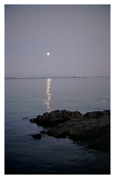 Color photograph of the bright moon over the Adriatic Sea - photographed from Volosko, Opatija. In the foreground, several rocks extend into the water from the right, and the light reflected by the moon creates a bright strip on the dark water. Towards the horizon, a container ship is anchored off the Istrian coast.