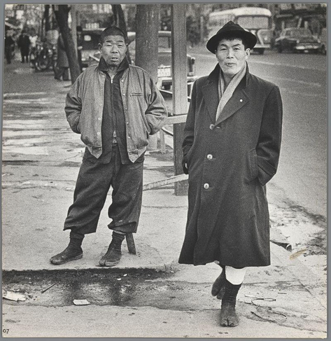 The image depicts two men standing on a city street, with one in the foreground and another slightly behind. The man closest to the viewer is dressed in a dark coat with large buttons and wears tall boots; he also sports a wide-brimmed hat that obscures his face from view. His expression appears composed but somber.

The second individual stands further back on what seems like a sidewalk or pedestrian area, donning casual attire consisting of a jacket over a shirt and trousers, accompanied by dark shoes. He has an unbuttoned posture with hands in pockets, suggesting nonchalance amid his surroundings.

Both subjects are set against the backdrop of urban life; buildings line one side of the street while another is partially visible on the right edge of the frame. There's evidence of daily activity: a vehicle parked further down the road and possibly some pedestrians or bystanders across from them. The environment appears to be that of an Asian city, likely indicative of Japan given historical context.

The overall tone rendered in grayscale adds gravity to this scene, reflecting perhaps on a bygone era where such street photography played a pivotal role documenting urban life during significant transitional periods within Japanese history, notably the post-war period mentioned alongside. The image carries with it not only visual content but also resonates as part of an archive that captures and immortalizes historical mome [...]