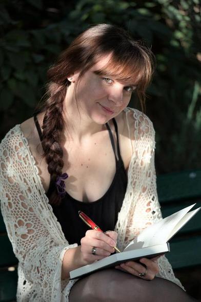 Une brune qui lève la tête de son carnet alors qu’elle y écrivait quelque chose pour regarder la caméra avec un léger sourire. Elle est éclairée par des tâche de soleil filtrant à travers des feuillages 