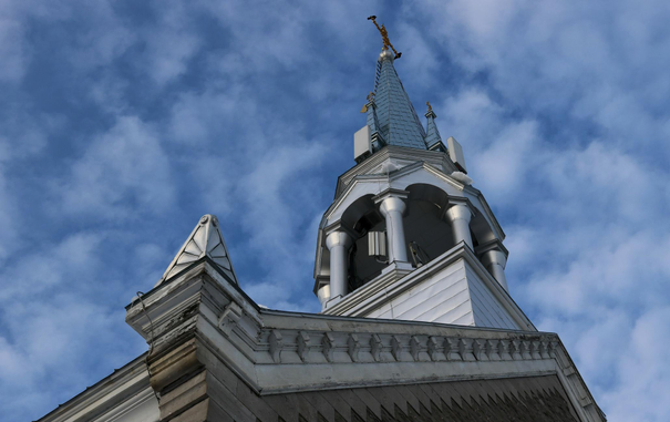 Photograph of a church steeple, taken looking upwards, near the corner of the building. In the foreground is a bent metal cornice, resembling a comical face with two eyes staring back at us. The roofline slopes steeply to the right, and the imposing, metal-clad steeple sits at the top centre of the image. At the tip of the pointed metal-shingled roof is a gilded cross, upon which two black crows are perched. The sky is a deep blue with small, fluffy clouds.

Photographie du clocher d'une église, prise en regardant vers le haut, tout près du coin du bâtiment. À l'avant-plan se trouve une corniche en métal plié, avec une apparence de visage comique avec deux yeux qui nous regardent. Le rebord du toit monte en angle vers la droite et le clocher imposant recouvert de placage en métal se trouve en haut au centre de la photo. Au bout du toit pointu en bardeaux de métal se trouve une croix dorée, où sont perchées deux corneilles noires. Le ciel est très bleu avec de petits nuages cotonneux.