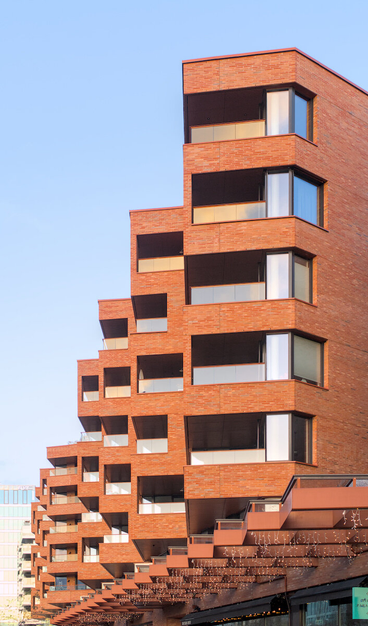 A modern apartment building in red brick, against a blue sky. The windows and balconies for a pattern that repeats _almost_ into the distance, with some variations that make it more interesting