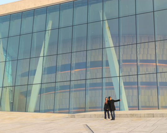 A close up photo of the facade of the Oslo Opera House. Usually there are crowds of tourists, but here there is only a couple. The man is looking to the side and pointing.