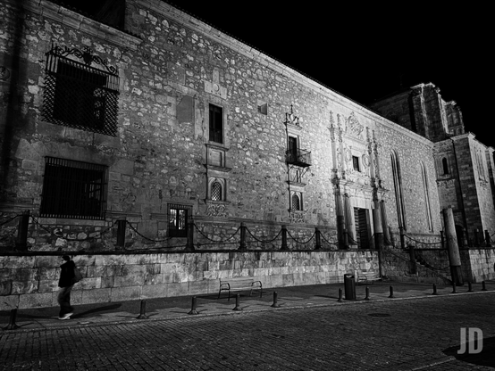 Imagen en blanco y negro de una escena nocturna, mostrando la fachada de un gran edificio antiguo de piedra, posiblemente una iglesia o monasterio, iluminado tenuemente. La pared de mampostería irregular presenta ventanas enrejadas y pequeños balcones de hierro forjado. En el centro, se distingue una entrada principal ornamentada con columnas. A la derecha, la estructura se eleva con elementos arquitectónicos más complejos, como contrafuertes o torres. En primer plano, una calle empedrada y una acera con bancos de metal y postes unidos por cadenas, donde una persona camina de izquierda a derecha, proyectando una sombra alargada.