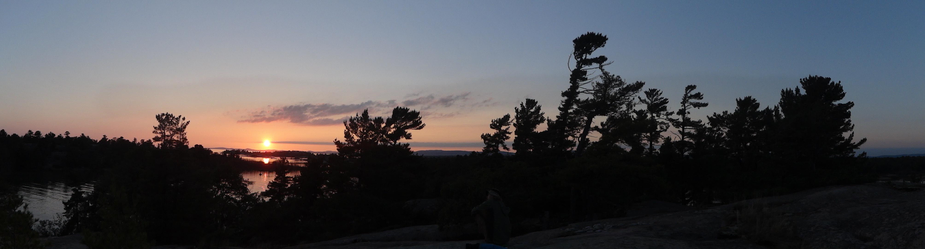 A panoramic photo taken from high up looking over the tops of pine trees in silhouette at a setting sun in the far distance over a distant shore with islands in the water between where the sun is setting and the tops of the trees in the foreground.