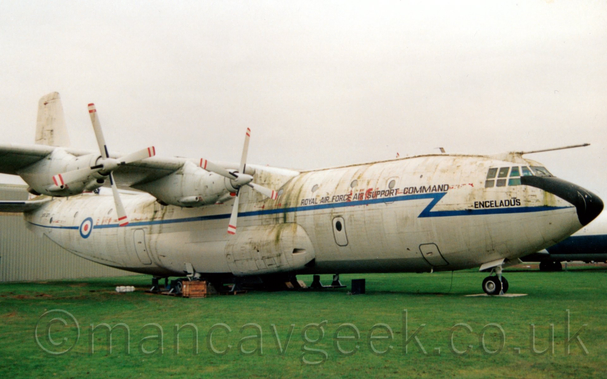 Side view of a high-winged, 4 propellor-engined military cargo aircraft parked on pads on grass, facing to the right.
The plane has a white upper half, the bottom being a light grey, with a blue stripe running along the body, zigzagging it's way through the forward fuselage.
There are black "Royal Air Force Air Support Command" titles on the upper forward fuselage, and the name "Enceladus" under the cockpit windows.
A large black panel covers the top of the nose, in front of the cockpit.
There is a blue, white, and red roundel overlaid on the blue stripe on the rear fuselage, and the black serial "XR371"  on the rear fuselage, under the tail.
The engines are suspended under and forward of the wing, with it's 4-bladed white propellors having red and white stripes on the tips.
Green grass fills the foreground at the bottom of the frame, with a grey building on the left of the frame behind the plane.
Flat grey sky fills the rest of the frame.