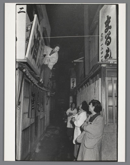 The image is a black and white photograph capturing an urban night scene, likely in Japan given the script on the signs. On the left side of the narrow alleyway stands a tall vending machine or payphone with Japanese characters above it marked "二" which translates to two. A person wearing traditional Japanese clothing, possibly for work based on their apron and demeanor, is perched atop this structure, seemingly interacting with its mechanism.

On the right side of the alleyway are three individuals dressed in light-colored coats, standing close together as if conversing or observing something intently. Their body language suggests attentiveness or curiosity towards the person above them.

The background features a building on the left displaying more Japanese characters and numbers "63 10" which could indicate an address or establishment number. The architecture of the buildings is simple, with wooden doors and louvered windows typical in traditional Japanese design.

This image appears to be part of a documentary series capturing various aspects of urban life during that period in Japan.