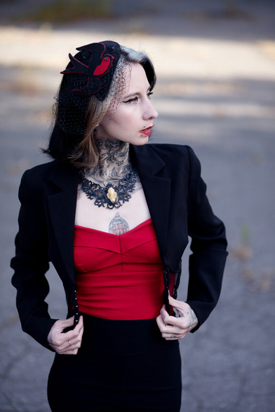 An alternative model's wide angle portrait. She is wearing a black and red outfit. Her neck's black tattoo and necklace reinforce the baroque tone of the portrait. Around her red lips, we can see a canine bite piercing.
