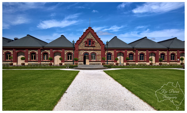 Full colour photograph of the main facade of Chateau Tanunda, featuring symmetrical red brick and stone work, arched windows, and dark sloping roofs.

A wide, bright green lawn and a central gravel pathway lead the eye toward the grand entrance under a bright blue sky with wispy white clouds.