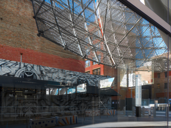 Reflection of street scene on a large glass pane, showing some of the curvy decorative structure and a brick building on the other side of the street.