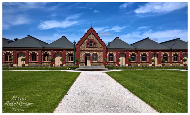 Full colour photograph of the main facade of Chateau Tanunda, featuring symmetrical red brick and stone work, arched windows, and dark sloping roofs.

A wide, bright green lawn and a central gravel pathway lead the eye toward the grand entrance under a bright blue sky with wispy white clouds.