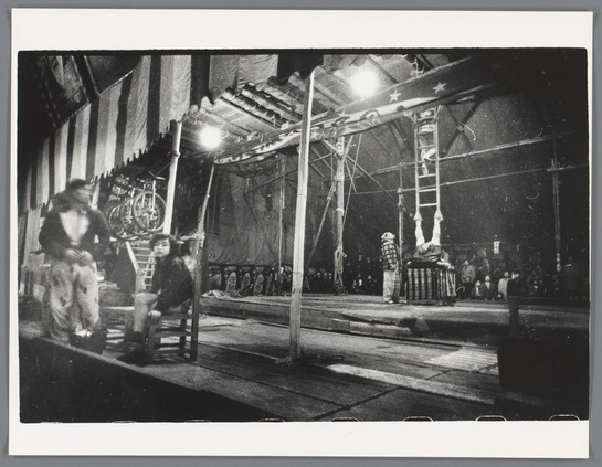 The image depicts a bustling circus scene with various performers and equipment. In the foreground, two individuals are seated on what appears to be part of a large wheel or contraption, possibly waiting for their turn to perform or simply observing. A third person is standing beside them in casual attire.

Behind this trio stands another individual dressed more formally, which suggests they might be overseeing operations or preparing for an act. Further back, the stage area reveals performers practicing or engaging with each other amidst circus paraphernalia such as ladders and props like a wheelbarrow on top of bicycles.

The overall setting is dimly lit by artificial lighting that highlights specific areas within this large tented structure known as a big-top. The dark background accentuates the monochromatic tones prevalent in black-and-white photography, capturing details from textures to forms with high contrast. This composition conveys both the energy and chaos associated with circus life while also showcasing individual expressions of anticipation or focus.

This photograph is notable for its vintage quality which indicates it was taken during an era when such circuses were a popular form of entertainment in Japan; specifically Osaka as indicated by the reference provided, suggesting that this image captures one moment within the broader historical context captured through van der Elsken's work on Japanese cultu [...]