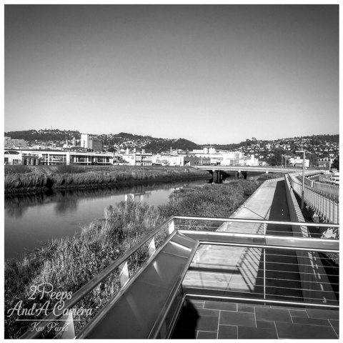 A wide, coloured landscape photograph showing a long, straight gravel path receding directly into the distance between two gently sloping grassy embankments.

The path appears to be on an elevated strip of land. In the background, the houses of Launceston stretch across the low hills under a bright blue sky. A large shadow is cast in the lower right corner.