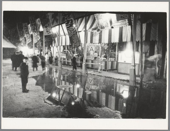 The image depicts a night-time scene at what appears to be an outdoor market or fair. Several large, striped tents and banners are illuminated by artificial light sources in the background, suggesting that they might be part of attractions such as rides or games typically found at fairs. A group of people is scattered throughout the foreground; some appear engaged with each other while others seem to be walking alone.

A notable feature is a puddle on the ground reflecting parts of the scene and creating distortions in perspective, possibly due to recent rainfall. The wet pavement suggests that it might have been raining prior or during this time when the photo was taken. Onlookers wear dark clothing which contrasts with the lighter tones of the tents.

The overall atmosphere conveys a sense of casual leisure activity among individuals who are partaking in or passing through what looks like a festive environment, possibly indicative of local culture and community gatherings around such events as fairs during cultural festivals. The image is monochromatic with black-and-white tonality enhancing its vintage feel which aligns well with the date specified in the reference information provided.

This photograph was taken by Ed van der Elsken based on his known work documented from a specific location, Osaka, Japan, and captures an environment that's characteristic of Japanese street scenes during festivals or local markets. T [...]
