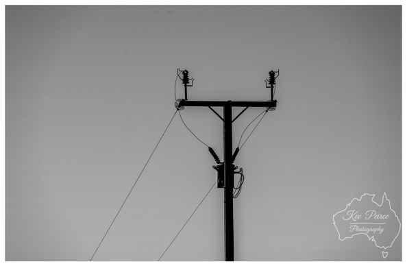 A high contrast, black and white photograph focusing on the top of a utility pole and its electrical lines, silhouetted against a smooth, bright gray sky.  The stark, geometric shapes of the insulators and transformer create a strong visual cross shape in the center of the frame, giving the image a graphic, minimalist feel. Photo by Kev Peirce.