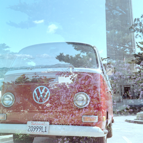 A double exposure color photo of a red Volkswagon Bus that is prominently taking up most of the frame under some blue skies with fluffy clouds with the Coit Tower in the background. Overlayed on it is another exposure of Alcatraz island in the distance, which is sitting right between the windshield and grill of the bus. The Alcatraz exposure has a some trees on the right that are faded in the sky and some purple flowers in the foreground which now looks painted onto the bus. Shot with a Rolleiflex Standard1935 and ZeissTessar75f3.5 lens, on KodakGold200, and developed with BelliniC41 by Shom Bandopadhaya. Licensed under Creative Commons Attribution-NonCommercial-ShareAlike (CC BY-NC-SA).
