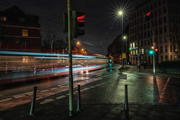 Das Bild zeigt eine Langzeitbelichtung, die vorbeifahrende Straßenbahn wird in dynamische Lichtspuren verwandelt und so einen Eindruck von Bewegung und Energie in der dunklen, nassen Umgebung vermittelt. Die Atmosphäre ist regennass und dunkel, akzentuiert durch helle Kunstbeleuchtung wie Laternen und Ampeln. Der Verkehr fließt, wie die roten und weiß-blauen Lichtstreifen des Fahrzeugs belegen. Die Szene ist von städtischen Gebäuden umgeben, darunter ein modernes Bürogebäude und ein älteres Gebäude mit traditionellerer Fassade.