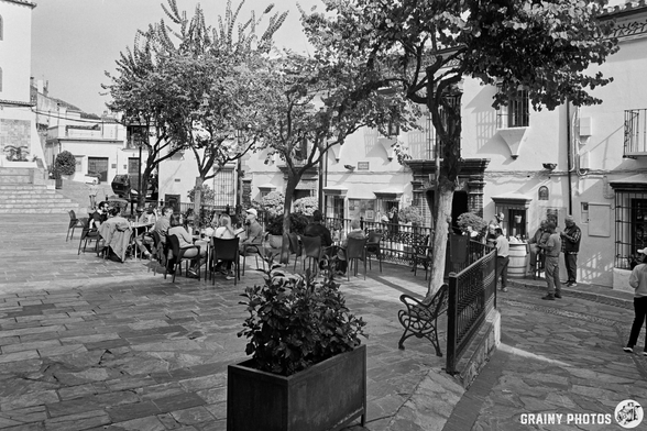 A bustling outdoor plaza surrounded by trees, featuring tables filled with diners enjoying their drinks and meals. In the background, charming white buildings and a few pedestrians create a lively atmosphere in this picturesque setting.