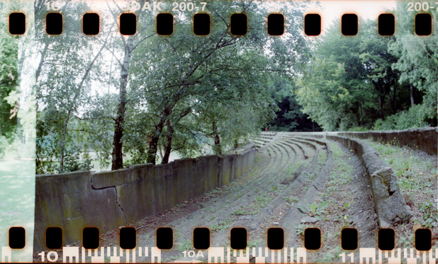 Detail einer alten Sportanlage. Von Grün überwachsen sieht man die steinerne Einfassung einer Tribüne. Die Sitzreihen folgen eine lang gestreckten Kurve und sind nach links durch eine Mauer begrenzt. Hinter der Mauer befindet sich die Schräge einer ehemaligen Radrennbahn. Die Betondecke ist von Birken aufgebrochen.