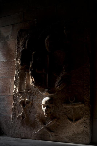 Une grande dalle de pierre rectangulaire très ancienne, avec des reliefs évoquant les Pirates, sûrement une pierre tombale, éclairée avec une lumière rasante depuis la gauche, donnant un air mystérieux et menaçant 