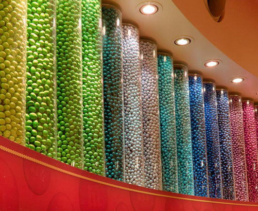 Un mur avec de gros tubes transparent, chacun remplis de M&Ms d’une même couleur, formant un dégradé de couleurs 