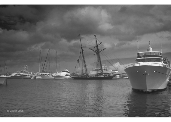A black and white image of a harbor with boats and yachts against a moody, cloudy sky. A majestic tall ship with sails is central, evoking nostalgia.