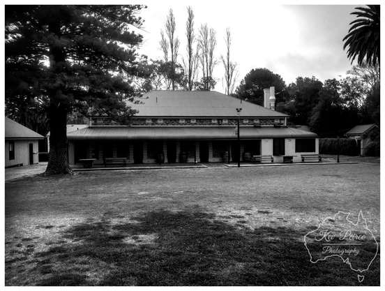 Black and white photograph of a long, historic stone building at the back of Seppeltsfield winery, featuring a wide verandah, a corrugated iron roof, and flanked by mature trees, with a large, shaded grassy lawn in the foreground.