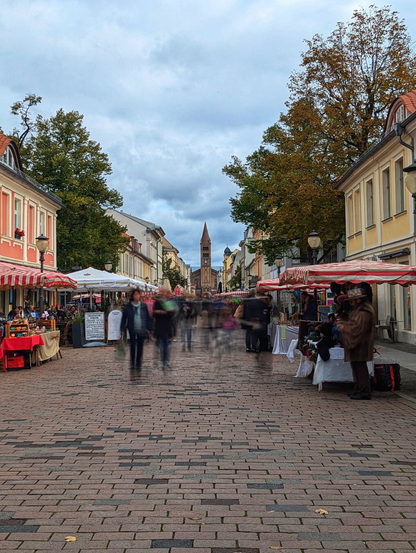 A bustling scene along Brandenburger Straße in Potsdam, featuring the iconic Church of St. Peter and Paul standing tall in the background. The cobblestone street is lined with pastel-coloured buildings, adding a quaint charm to the atmosphere. Market stalls and outdoor seating areas, adorned with red-and-white striped awnings and white umbrellas, create a lively and inviting environment. People are seen walking and browsing, their movement artistically blurred, capturing the dynamic energy of the street. Autumnal trees frame the scene, while an overcast sky lends a soft, diffused light to the picturesque setting.
