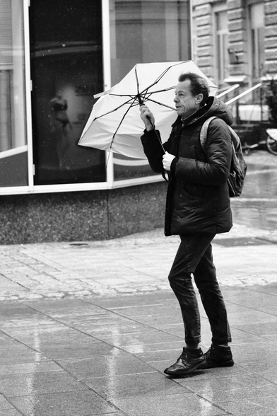 Das Foto zeigt eine Straßenszene in Schwarz-Weiß in der St. Pöltenter Innenstadt an einen regnerischen windigen Tag. Man sieht eine Person mit Regenschirm der durch den Wind verbogen ist.